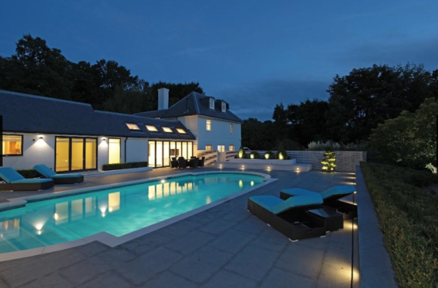 A modern house features a serene swimming pool surrounded by lounge chairs. The scene is set during twilight, highlighting the elegant design and lush greenery in the background.