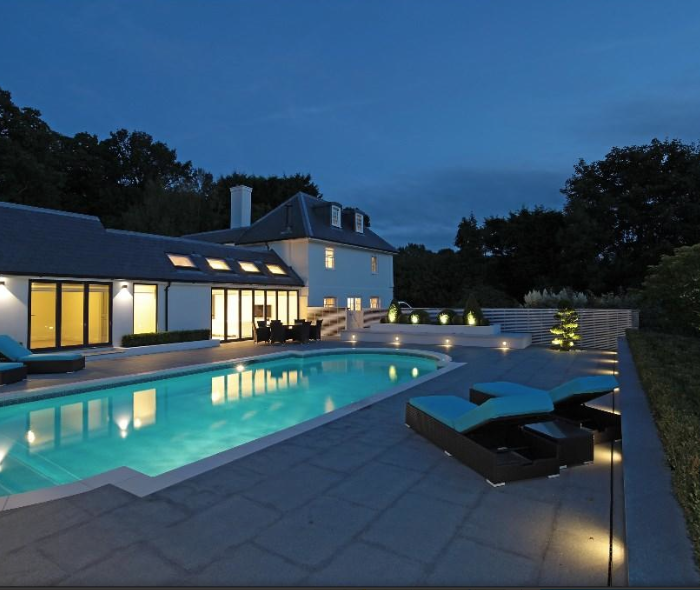 A modern house features a serene swimming pool surrounded by lounge chairs. The scene is set during twilight, highlighting the elegant design and lush greenery in the background.