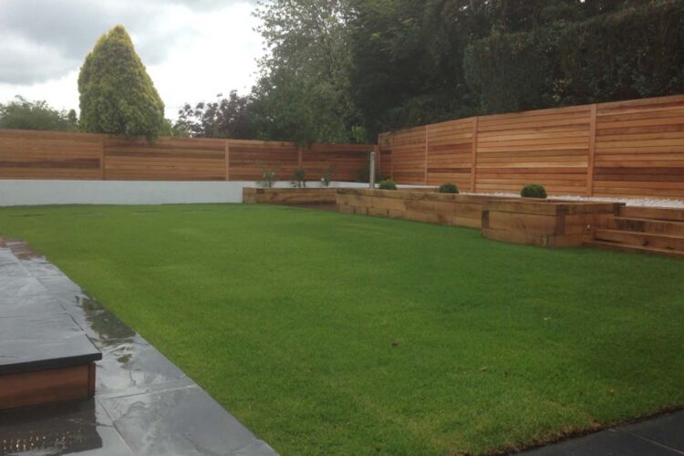 A green lawn occupies the foreground, with wooden planters and a gravel area in the background. A wooden fence provides privacy, and trees border the space under an overcast sky.