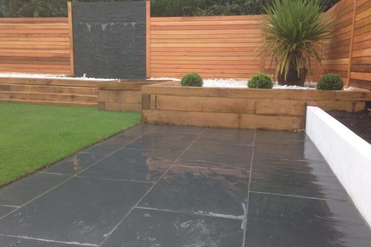 A slate patio features a small water fountain against a dark stone wall, surrounded by wooden fencing. Lush green grass and neatly trimmed shrubbery enhance the serene outdoor setting.