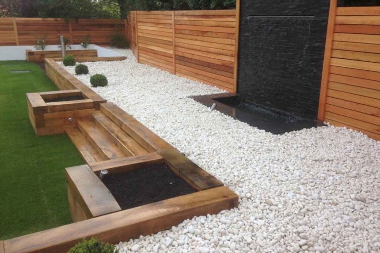 A minimalist garden features wooden planters and steps surrounded by white gravel. A water feature cascades down a black wall, with green shrubs adding a touch of greenery. Wooden fences provide enclosure.
