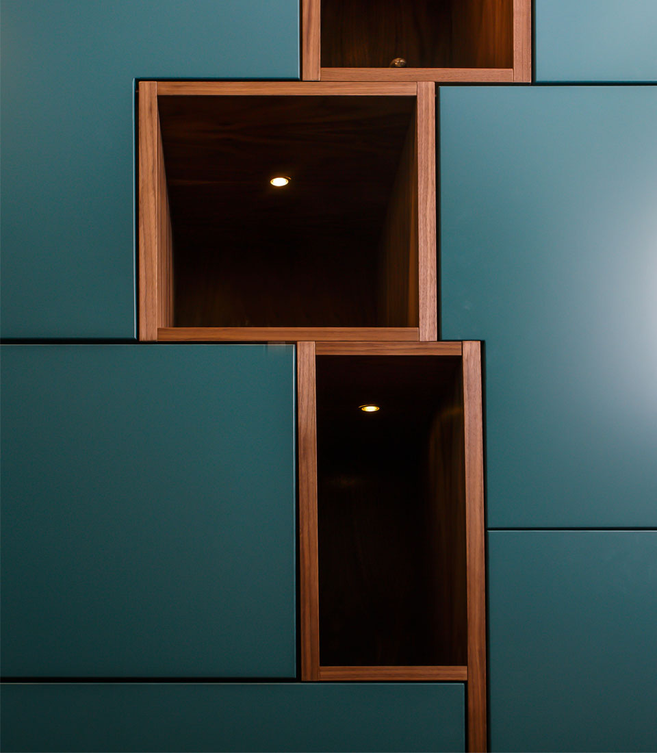 A series of rectangular wooden shelves, inlaid within a teal wall, feature warm lighting inside some spaces, creating an elegant contrast in the modern interior design.