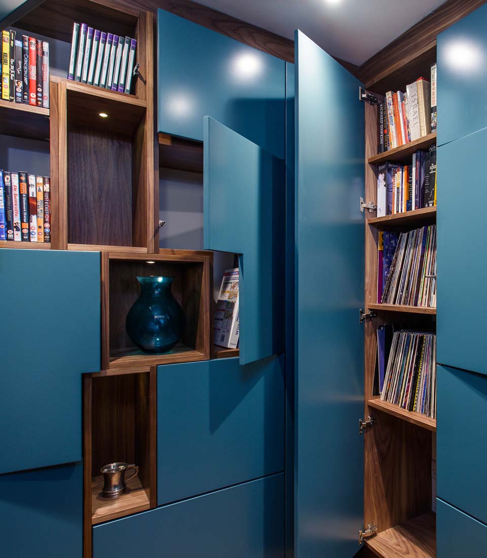 A modern bookshelf features teal doors, open shelves, and wooden cabinetry. Inside, DVDs, books, vinyl records, and decorative items like a blue vase and a small silver pitcher are organized.