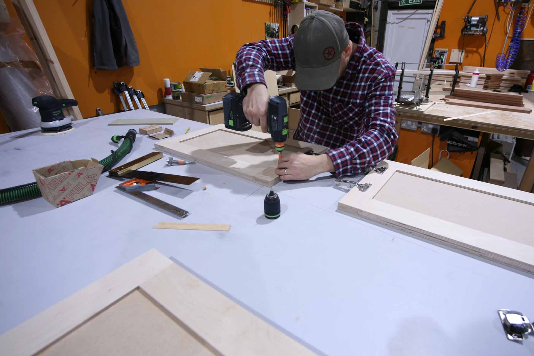 A person in a plaid shirt uses a power drill to assemble wood pieces on a worktable cluttered with tools, with an orange wall and organized storage in the background.