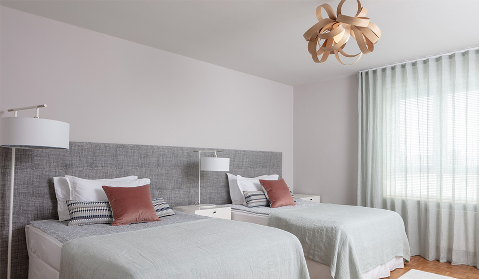 Two neatly arranged beds with light green bedspreads and decorative pillows occupy a room. A modern light fixture hangs above, and sheer curtains filter natural light from a window.