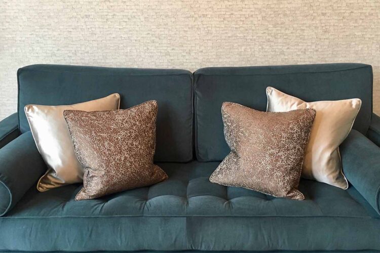 A plush teal couch features two shiny silver and patterned brown decorative pillows, positioned on a tufted seat, set against a textured, light-colored wall.
