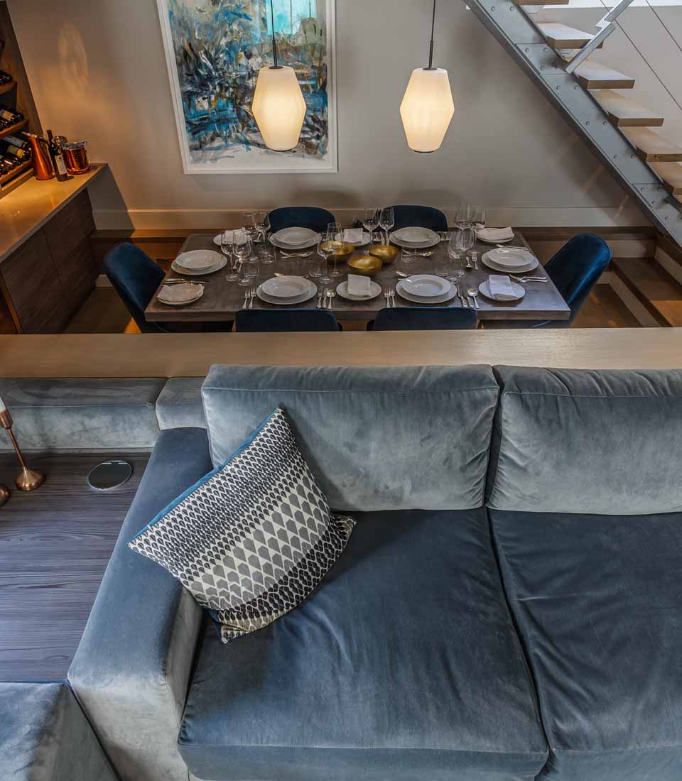 A plush blue couch with a patterned pillow sits next to a dining area featuring a table set for a meal, illuminated by hanging lights in a modern interior space.