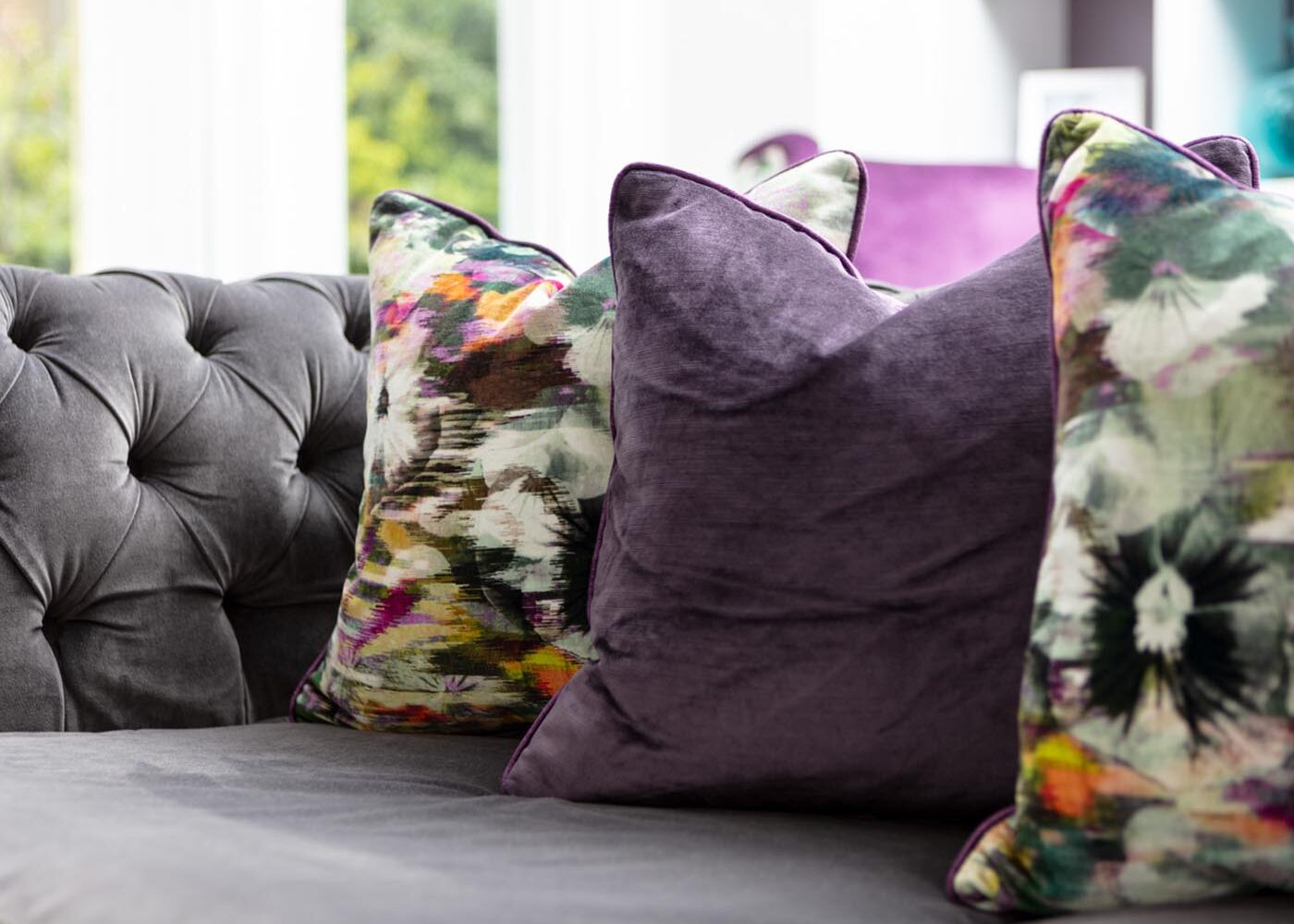 A grey tufted sofa features three decorative pillows: two are purple and one is vibrant with floral patterns, set against a light-filled modern interior.