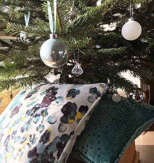 A decorated Christmas tree with hanging ornaments stands above two decorative pillows, one featuring a floral pattern and the other a textured design, placed on a wooden surface.