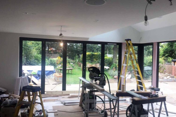 A room under renovation features construction tools, a tall ladder, and a table saw. Large glass doors open to a garden with greenery and furniture visible outside.