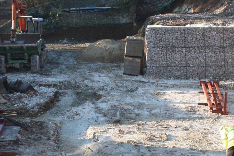 A green construction vehicle is loading material in a gravelly site, surrounded by piles of stones and dirt. In the background, a stone wall is being built, and workers are present.