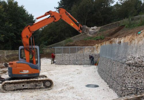 An excavator scoops gravel while two workers construct a stone wall in a gravel pit. Surrounded by trees, the site is partially cleared with visible dirt mounds and tools.
