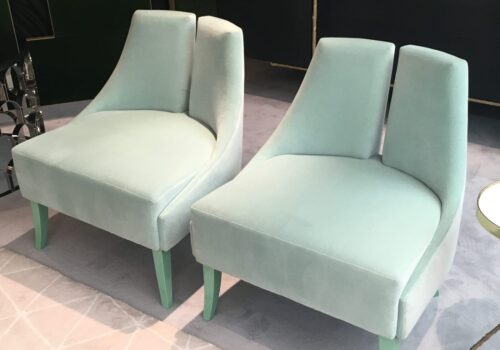 Two elegant, mint green velvet chairs sit closely together, featuring smooth curves and light green legs, on a soft, light gray patterned rug, in a stylish, contemporary setting.
