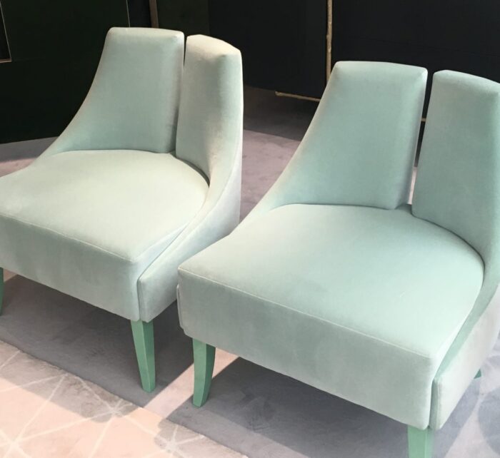Two elegant, mint green velvet chairs sit closely together, featuring smooth curves and light green legs, on a soft, light gray patterned rug, in a stylish, contemporary setting.