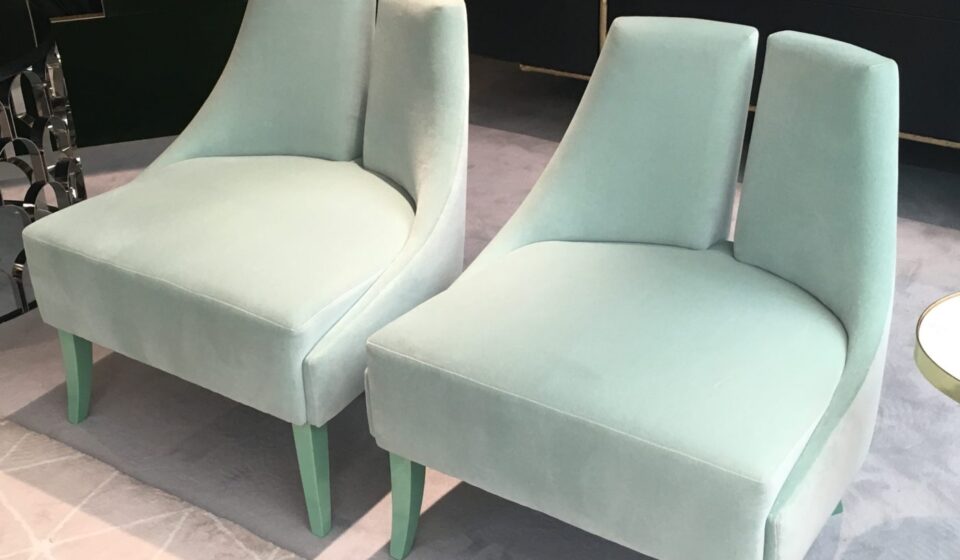 Two elegant, mint green velvet chairs sit closely together, featuring smooth curves and light green legs, on a soft, light gray patterned rug, in a stylish, contemporary setting.