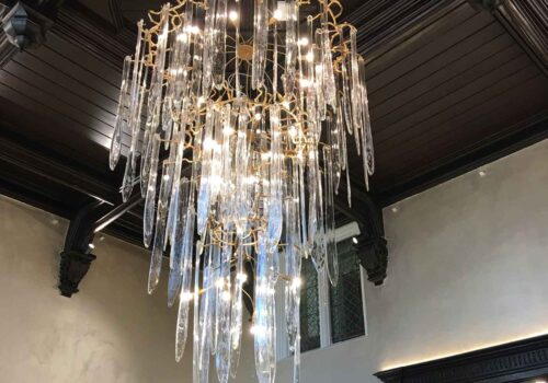 A luxurious chandelier with elongated glass pieces hangs from a dark wooden ceiling, illuminating the surrounding elegant room while contrasting with the light-colored walls.