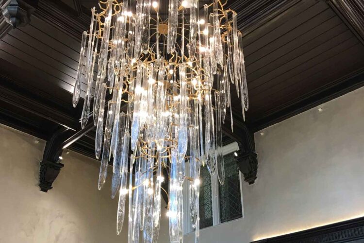 A luxurious chandelier with elongated glass pieces hangs from a dark wooden ceiling, illuminating the surrounding elegant room while contrasting with the light-colored walls.