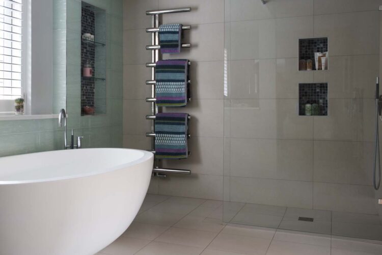 A modern bathroom features a freestanding white bathtub, a glass shower, and a towel rack with colorful towels. The walls are tiled in soft colors, creating a serene atmosphere.