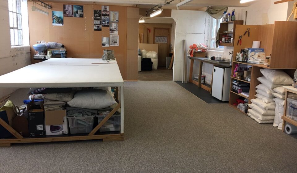 A spacious workroom features a large white table and cabinets filled with supplies. Shelves hold stacked materials, while a kitchenette is visible in the background. The floor is carpeted.