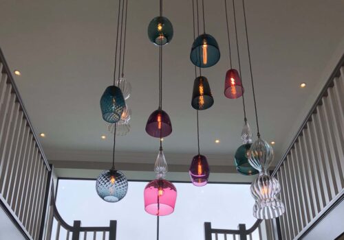 Colorful glass pendant lights hang from the ceiling, illuminating a modern staircase in a spacious interior, with large windows providing natural light and a view outside.