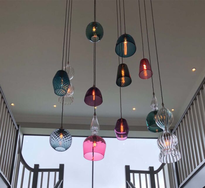Colorful glass pendant lights hang from the ceiling, illuminating a modern staircase in a spacious interior, with large windows providing natural light and a view outside.