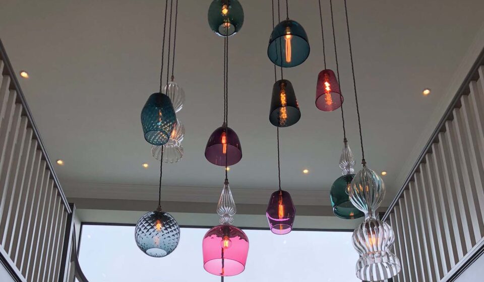 Colorful glass pendant lights hang from the ceiling, illuminating a modern staircase in a spacious interior, with large windows providing natural light and a view outside.