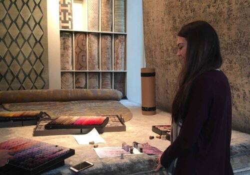 A woman examines color samples on a work table, surrounded by rolls of textured fabric and rugs on display. The environment suggests a design studio or showroom.