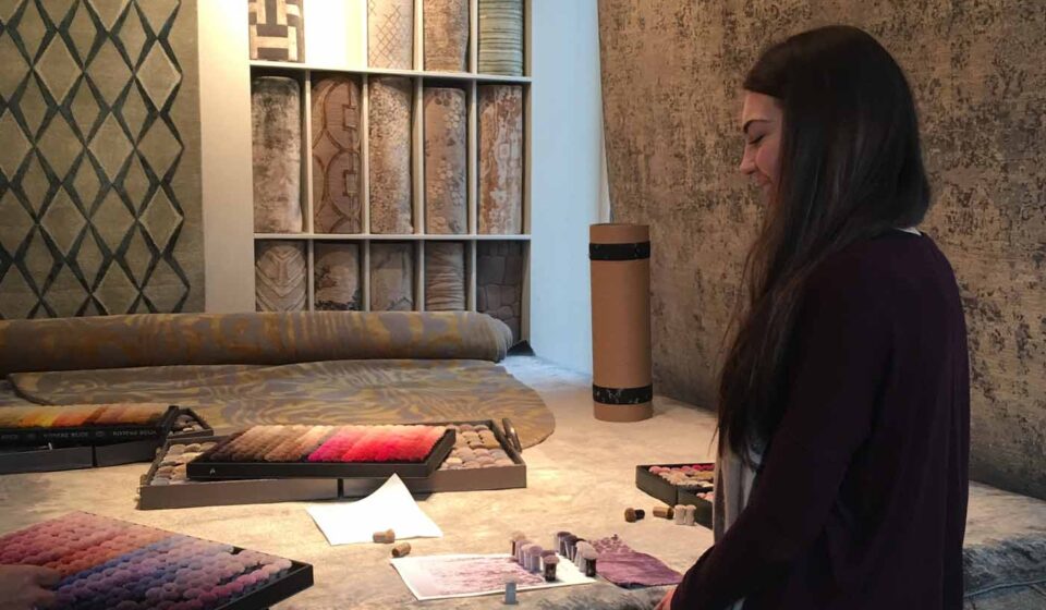 A woman examines color samples on a work table, surrounded by rolls of textured fabric and rugs on display. The environment suggests a design studio or showroom.