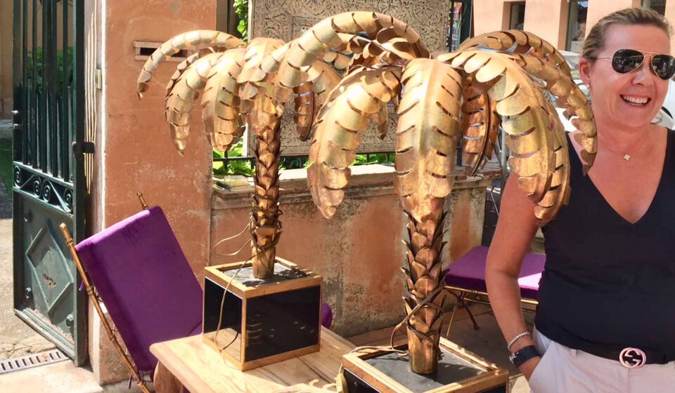 Two golden palm tree sculptures stand on a table beside a smiling woman. A purple folding chair leans against the table, set against a sunny, vibrant outdoor setting.