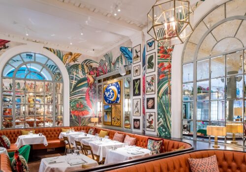 A stylish restaurant features a vibrant mural of tropical plants and animals. Under warm lighting, round tables are set with white tablecloths and glassware, complementing the plush, brown banquettes.