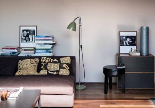 A brown sofa with patterned pillows sits next to a stack of books and a green lamp. A small black stool and a dark wooden cabinet display framed photos and decorative items in a minimalist setting.