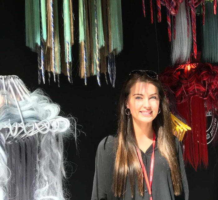 A young woman stands smiling next to a collection of colorful hanging lamps made from various materials, against a dark backdrop. Sunlight casts shadows across her face and surroundings.
