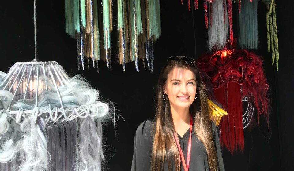 A young woman stands smiling next to a collection of colorful hanging lamps made from various materials, against a dark backdrop. Sunlight casts shadows across her face and surroundings.