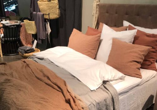 A neatly made bed features various textured linens in neutral and warm tones, with multiple cushions arranged attractively. Nearby, a dining table with glasses and woven baskets is visible in a styled room.