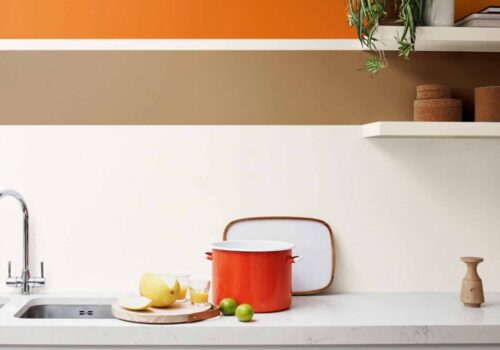 A bright red pot sits on a white countertop next to two limes and a yellow fruit, with a sleek faucet and abstract color-blocked wall above, featuring orange and brown stripes.
