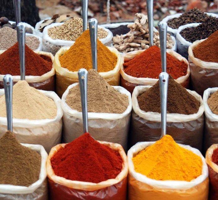Numerous bags filled with various colored spices sit in a market setting. Each bag has a metal scoop on top, showcasing a vibrant array of textures and hues.