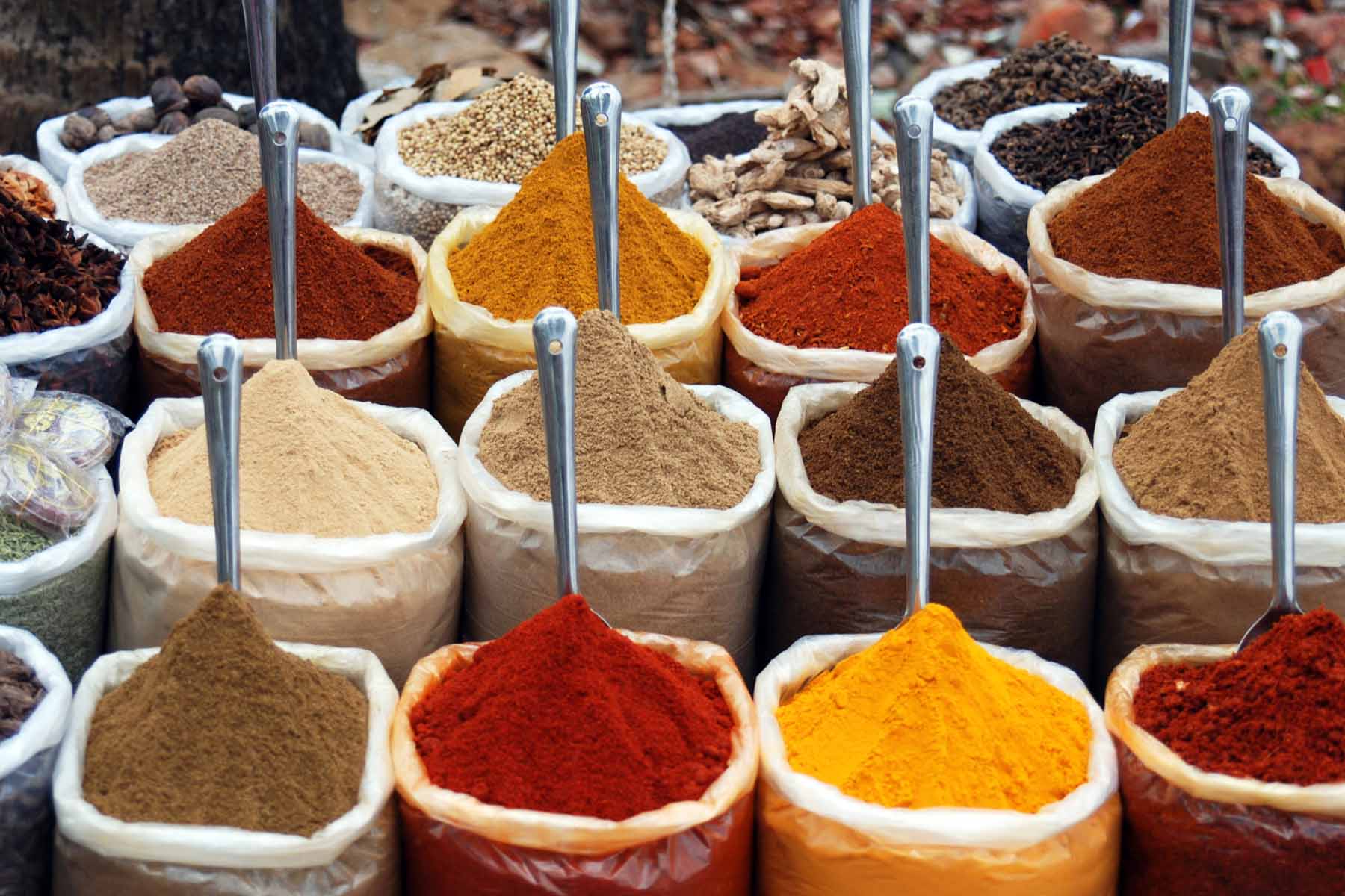 Numerous bags filled with various colored spices sit in a market setting. Each bag has a metal scoop on top, showcasing a vibrant array of textures and hues.