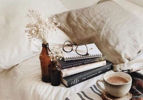 A stack of books rests on a bed, accompanied by a coffee cup and a small vase with dried flowers. A pair of glasses sits atop the books, nestled against soft pillows.