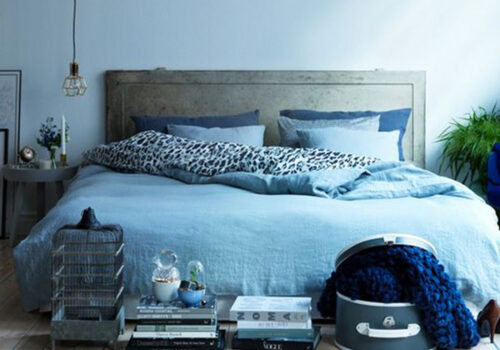 A bed adorned with blue linens and patterned pillows occupies the center. Nearby, a table holds flowers and candles. A caged decoration and stacked books add charm, while a green plant enhances the atmosphere.