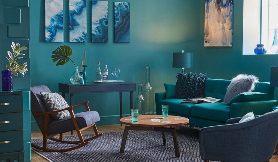 A turquoise living room features a cozy sofa with cushions and a shaggy throw, a wooden coffee table with glasses, and a rocking chair. Art and vases adorn the walls.