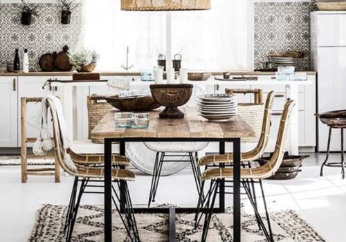 A wooden dining table sits center stage, surrounded by chairs. On the table, a large bowl holds decorative items. The bright kitchen features patterned tiles and light curtains, enhancing the ambiance.