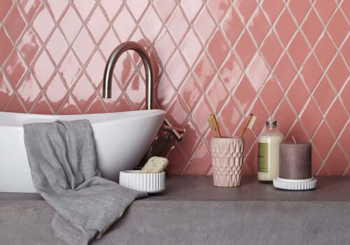 A white freestanding bathtub rests against a pink diamond-patterned wall. A gray towel drapes over the side, while various toiletries and candles are arranged on a concrete surface nearby.