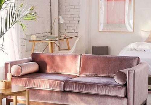 A plush pink sofa with rounded armrests is centrally placed in a bright, airy room. A desk and chair are nearby, with a plant adding a touch of greenery.