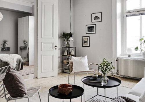 A cozy living room features a white armchair, black circular tables with plants, and a white door leading to a bedroom. Natural light floods in through a window, highlighting minimalist decor.
