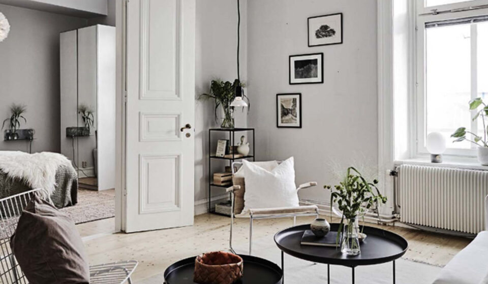 A cozy living room features a white armchair, black circular tables with plants, and a white door leading to a bedroom. Natural light floods in through a window, highlighting minimalist decor.