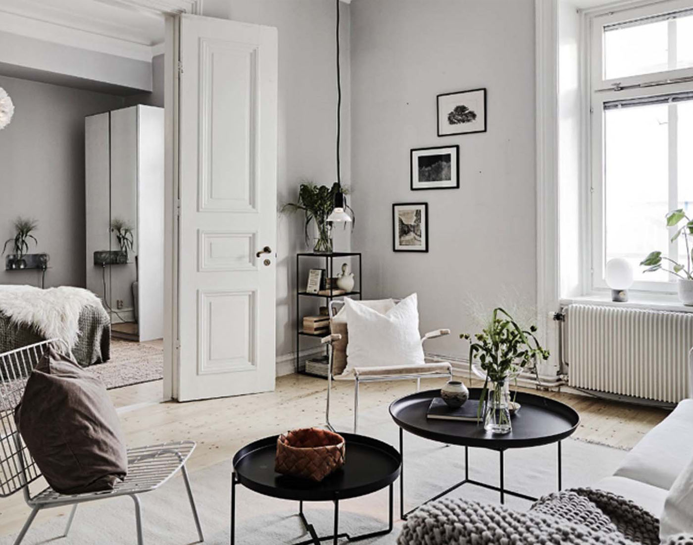 A cozy living room features a white armchair, black circular tables with plants, and a white door leading to a bedroom. Natural light floods in through a window, highlighting minimalist decor.