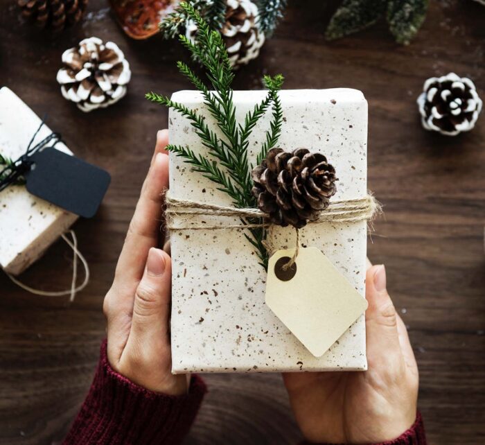 A person holds a wrapped gift decorated with a pinecone and sprigs on top. Nearby, festive elements include other gifts, pinecones, and a cup with a snowman-shaped marshmallow on a wooden table.