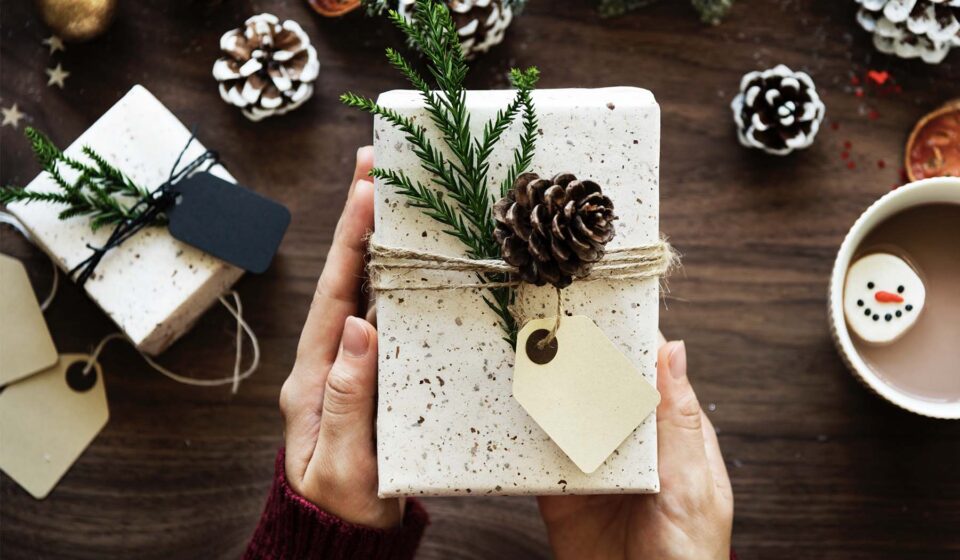 A person holds a wrapped gift decorated with a pinecone and sprigs on top. Nearby, festive elements include other gifts, pinecones, and a cup with a snowman-shaped marshmallow on a wooden table.