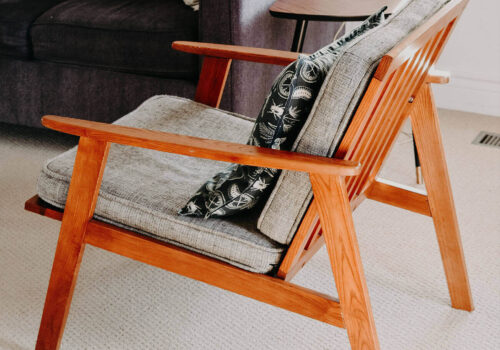 A wooden armchair with gray cushions and a decorative pillow sits in a cozy living room, beside a small, round table and a dark sofa, on a light carpeted floor.