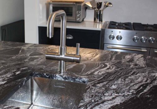 A sleek, silver kitchen faucet is positioned above a rectangular stainless steel sink, which sits on a dark, patterned countertop. In the background, a stove and microwave are visible, suggesting a modern kitchen setting.
